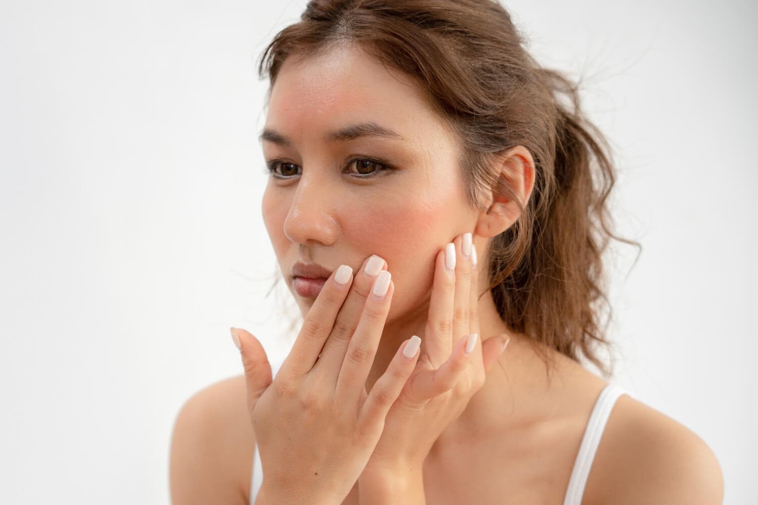 Woman in a white tank top touching her cheek with both hands, showing redness across her cheeks and forehead. Woman in a white tank top touching her cheek with both hands, showing redness across her cheeks and forehead.