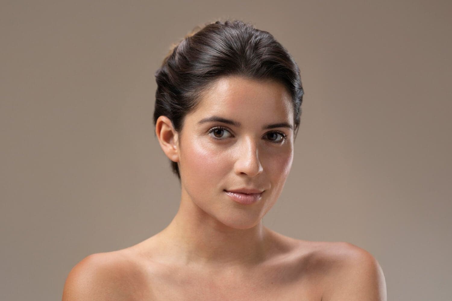 Bare-shouldered woman with dark hair swept back, wearing dewy bare skin with only a soft flush and glossy lip, against a taupe backdrop. Bare-shouldered woman with dark hair swept back, wearing dewy bare skin with only a soft flush and glossy lip, against a taupe backdrop.