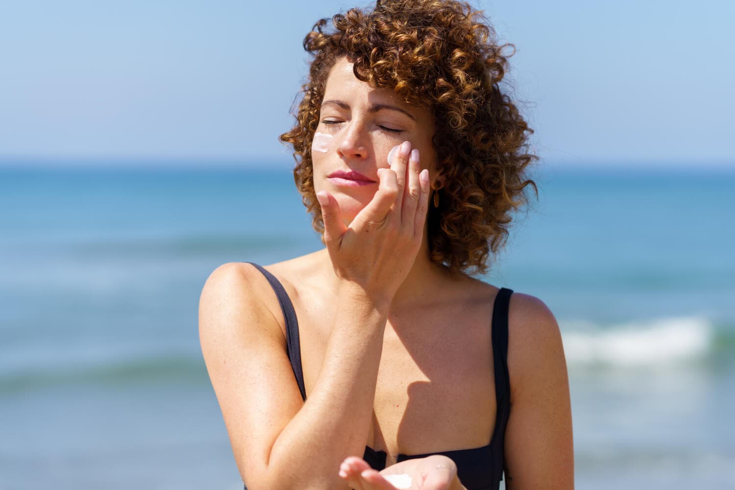 Curly-haired woman in a black swimsuit blending a white streak of sunscreen into her cheek on a sunlit beach, sea behind her.