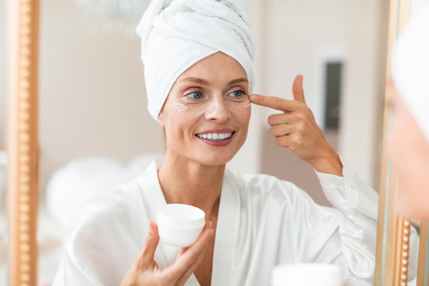 Smiling woman in a white robe and hair towel patting cream under her eye while holding an unlabelled white jar at the mirror. Smiling woman in a white robe and hair towel patting cream under her eye while holding an unlabelled white jar at the mirror.