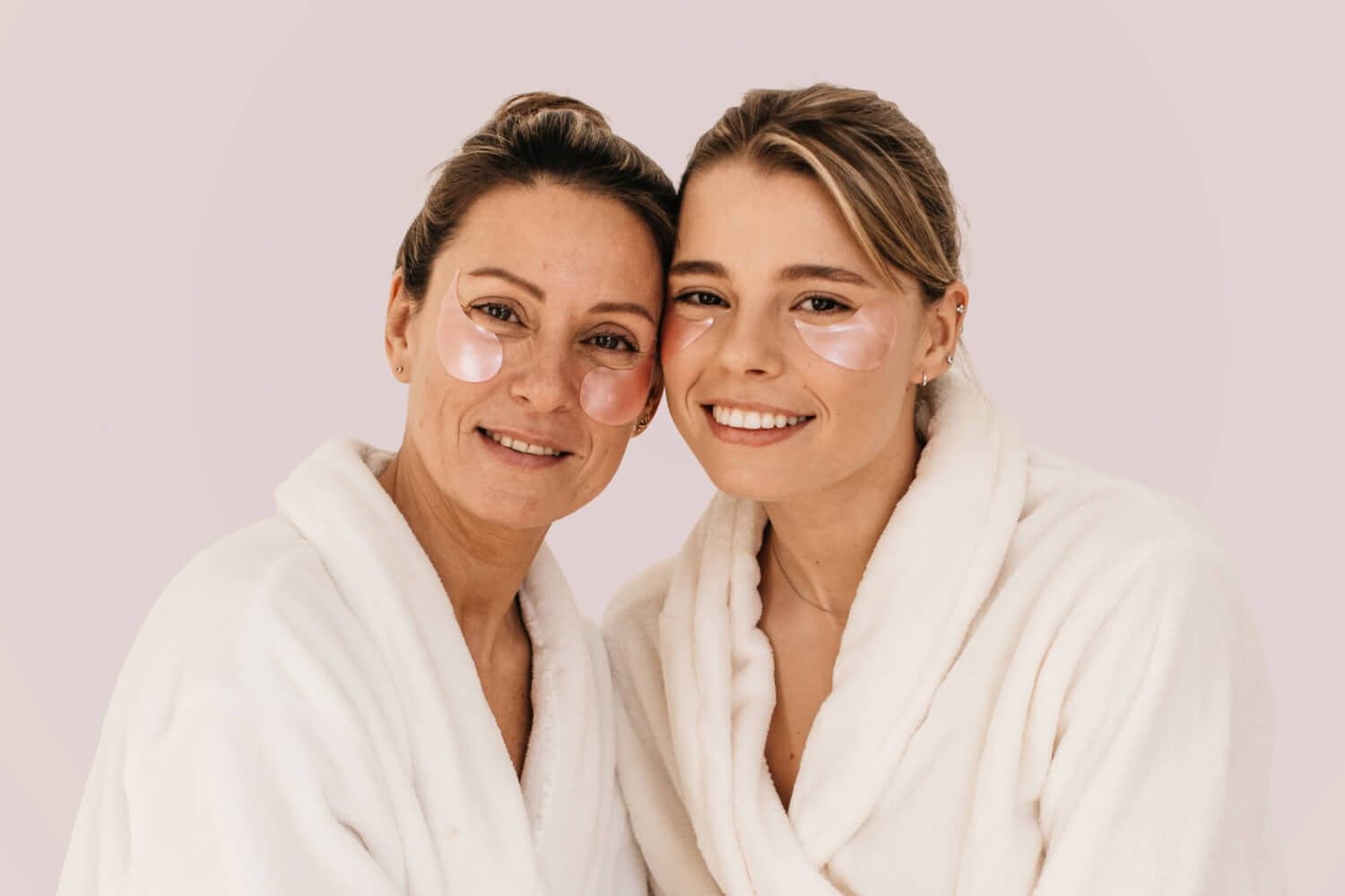 Two smiling women in white towelling robes lean cheek to cheek, both wearing pink under-eye gel patches, against a soft lilac backdrop. Two smiling women in white towelling robes lean cheek to cheek, both wearing pink under-eye gel patches, against a soft lilac backdrop.