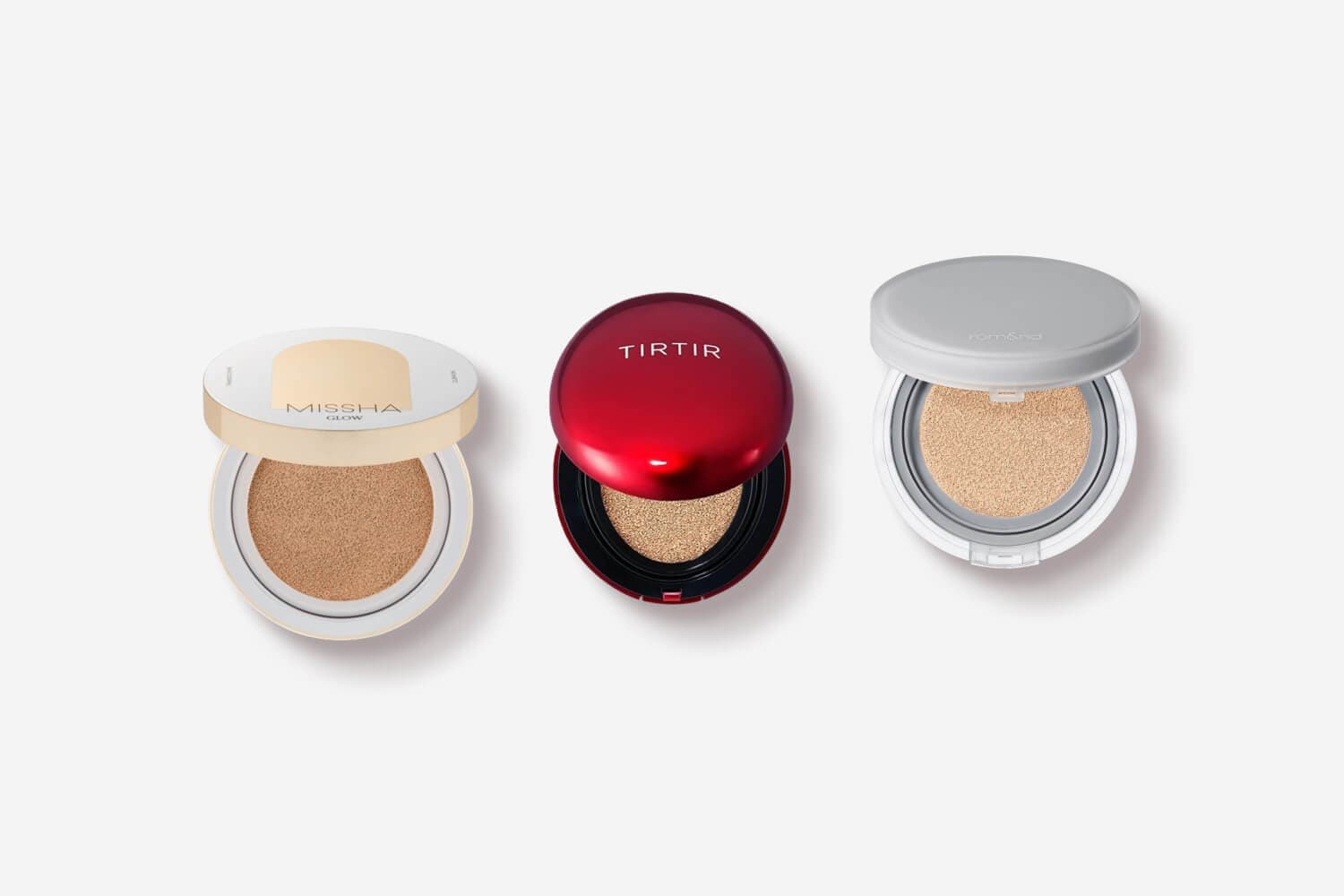 Three open cushion compacts on a grey background: Missha Glow in cream and gold, TIRTIR in red, and rom&nd in white and grey. Three open cushion compacts on a grey background: Missha Glow in cream and gold, TIRTIR in red, and rom&nd in white and grey.