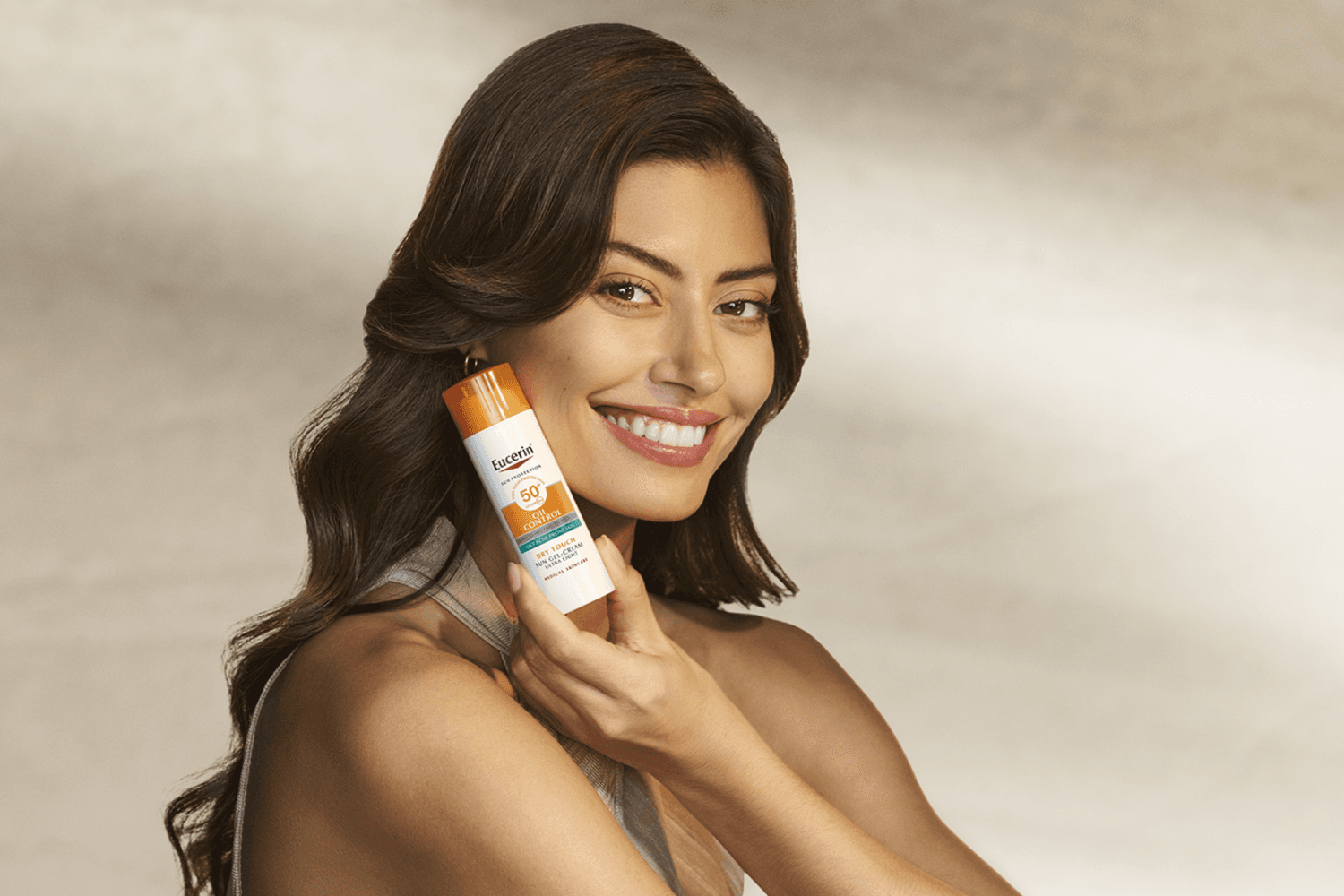 Smiling dark-haired woman holding a bottle of Eucerin Sun Protection Oil Control Dry Touch Sun Gel-Cream SPF 50 beside her cheek. Smiling dark-haired woman holding a bottle of Eucerin Sun Protection Oil Control Dry Touch Sun Gel-Cream SPF 50 beside her cheek.
